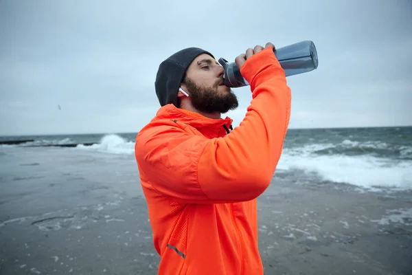 Close-up of young attractive dark haired bearded runner dressed in warm athletic clothes holding fitness bottle in raised hand while drinking water after morning workout — Stock Photo, Image