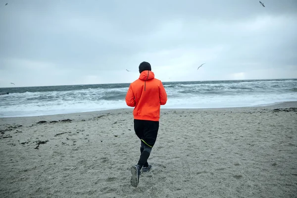 Achteraanzicht van fitness mannelijk model trainen en lopen aan zee op grijze vroege ochtend, gekleed in warme sportieve kleding en oranje jas. Sport en gezonde levensstijl — Stockfoto