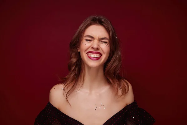 Agradável olhar alegre adorável jovem mulher cabelos castanhos com maquiagem à noite sorrindo amplamente com os olhos fechados, posando sobre fundo claret com confete de prata em sua cabeça — Fotografia de Stock