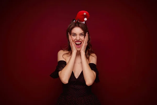 Ευτυχισμένη ελκυστική νεαρή καστανή κυρία φορώντας κομψό φόρεμα με κόκκινες τελείες και καπέλο santa ποζάροντας πάνω από το κόκκινο φόντο, χαμογελώντας ευρέως στην κάμερα με ενθουσιασμένο πρόσωπο — Φωτογραφία Αρχείου