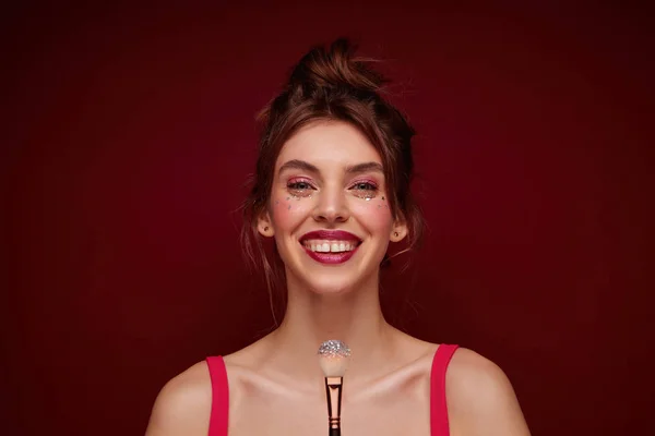 Close-up of positive young brunette woman with bun hairstyle smiliing happily at camera and holding brush with lushes, making evening makeup while posing over burgundy background — Stok Foto