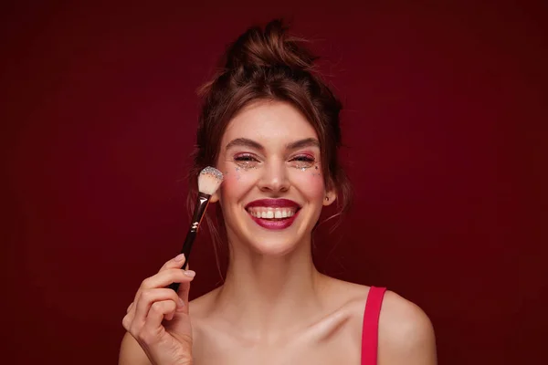 Happy young brown haired lovely woman with festive makeup wearing casual hairstyle while standing over burgundy background, meeting with friends in evening and being in high spirit