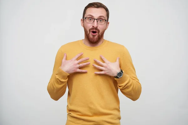 Open-eyed pretty young bearded guy in eyewear with brown short hair holding palms on his chest and looking surprisedly at camera, isolated over white background — Stock Photo, Image