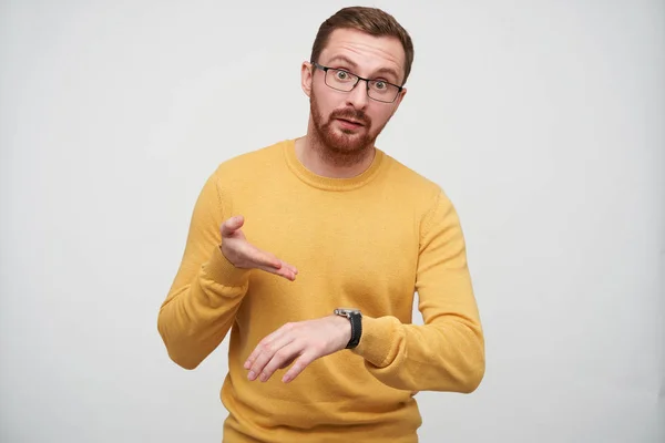 Dissatisfied young brown haired bearded guy in eyewear pointing indignantly on his with wristlet watch, being discontented that someone is late, isolated over white background — Stock Photo, Image