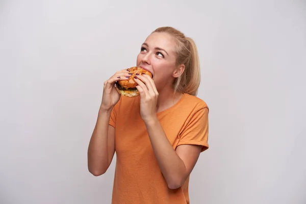 Indoor foto van tevreden jonge langharige blonde vrouw met paardenstaart kapsel dragen casual kleding terwijl poseren over witte achtergrond, het eten van smakelijke hamburger en kijken positief opzij — Stockfoto