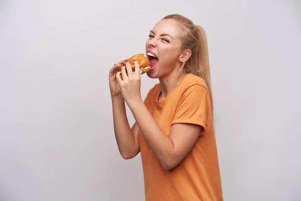 Studio shot van opgewonden jonge mooie blonde dame met casual kapsel houden hamburger in haar handen en gaan afbijten stuk van het, op zoek vraatzuchtig naar camera met brede mond geopend — Stockfoto