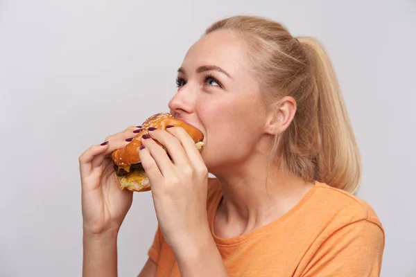 Close-up van jonge mooie positieve blonde vrouw met casual kapsel verrukkelijke frisse smakelijke hanburger staan over witte achtergrond in casual t-shirt — Stockfoto