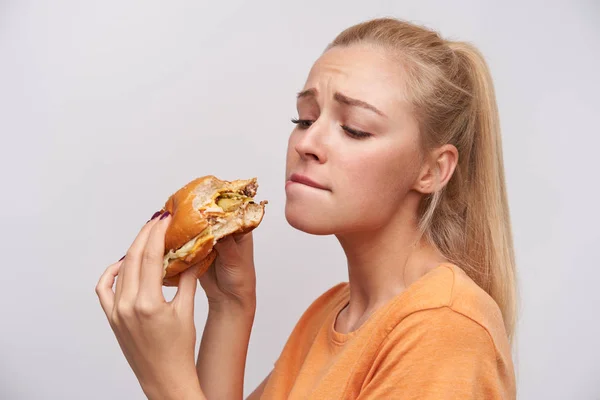 Portret van jonge mooie langharige blonde vrouw met paardenstaart kapsel houden hamburger in opgeheven handen en kijken onverzadigbaar op, bijten onderlip en fronsende wenkbrauwen over witte achtergrond — Stockfoto