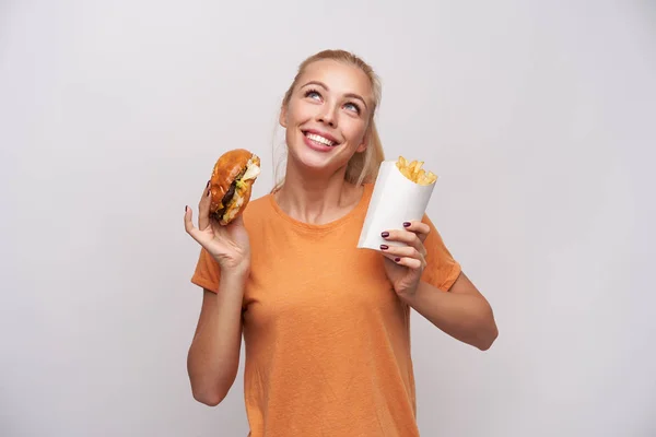 Vrolijke aantrekkelijke jonge blauw-ogige blonde vrouw met hamburger en frietjes in opgeheven handen en gelukkig opwaarts kijkend, lachend op grote schaal terwijl ze over een witte achtergrond poseert — Stockfoto