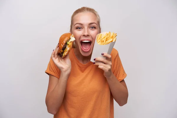 Overgelukkig jonge blauw-ogige blonde vrouw met casual kapsel kijken opgewonden op camera met brede ogen en mond geopend, met hamburger en frietjes terwijl staan over witte achtergrond — Stockfoto