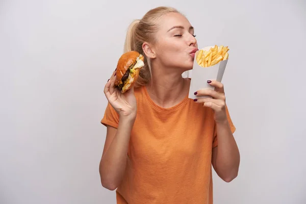 Aangename goed uitziende jonge mooie blonde vrouw in casual kleding staan over witte achtergrond met fast food in opgeheven handen, het houden van de ogen gesloten tijdens het voorspellen van lekker eten — Stockfoto