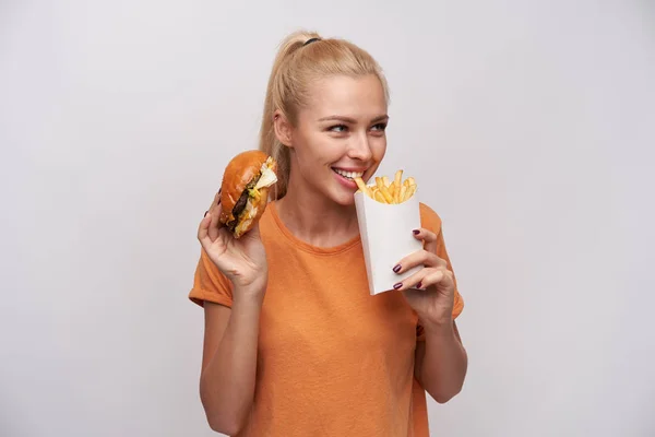Portret van mooie jonge blonde vrouw met paardenstaart kapsel eten frites en kijken positief opzij, glimlachen op grote schaal terwijl staan over witte achtergrond — Stockfoto