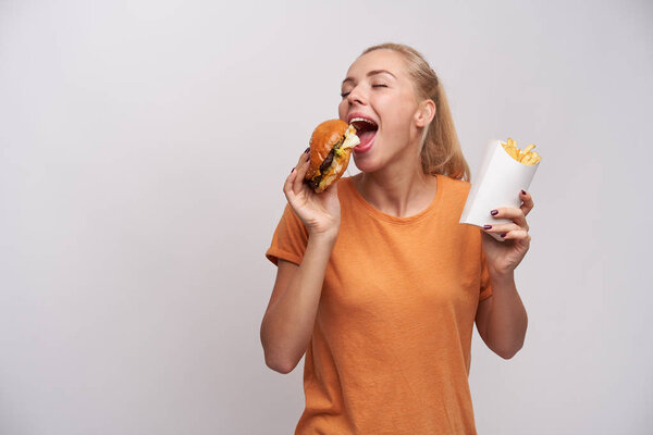 Pleasant looking positive young blonde lady with ponytail hairstyle keeping eyes closed while enjoying her tasty burger, standing over white background in casual clothes