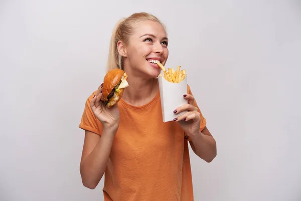 Indoor foto van aantrekkelijke jonge blonde dame in oranje t-shirt glimlachen vrolijk tijdens het trekken met tanden frites en houden hamburger in andere hand, geïsoleerd over witte achtergrond — Stockfoto