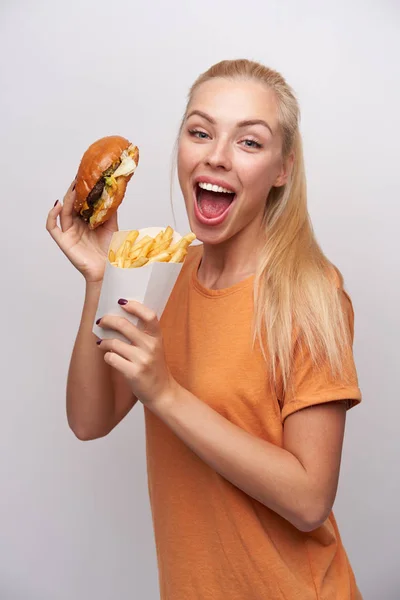 Mulher loira de cabelos compridos jovem super alegre com fastfood em mãos levantadas olhando feliz para a câmera com olhos largos e boca aberta, isolado sobre fundo branco — Fotografia de Stock