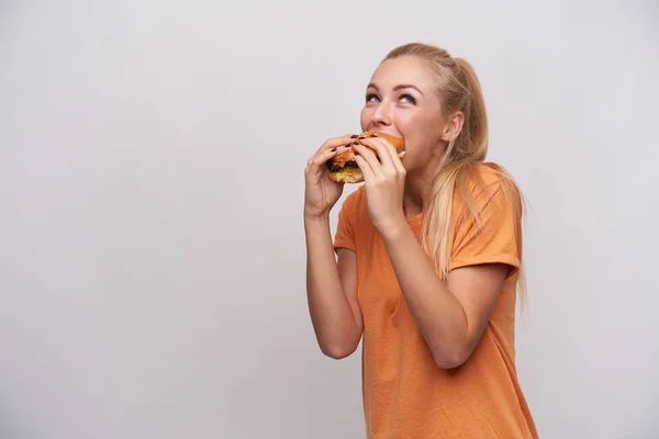 Indoor photo of young cheerful blonde female with casual hairstyle eating hamburger with great pleasure while posing over white background in orange t-shirt — Stock Photo, Image