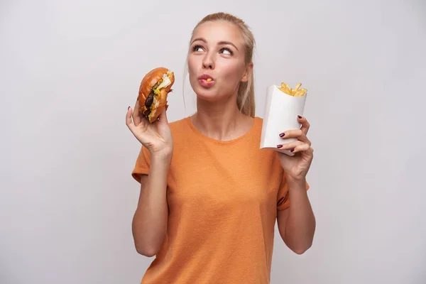 Studio photo of young long haired blonde lady holding junk food in raised hands and looking positively upwards, wearing casual clothes while posing over white background — Stock Photo, Image