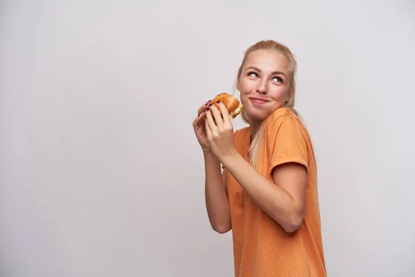 Studio shot van hongerige jonge blonde vrouw in oranje t-shirt kijken positief opzij met mond vol eten, het houden van verse hamburger terwijl staande over witte achtergrond — Stockfoto