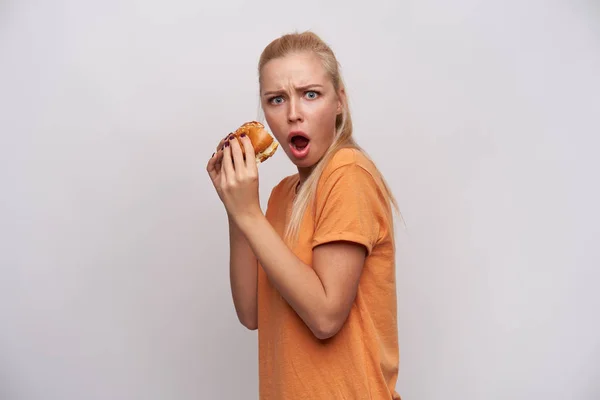 Scioccato giovane attraente donna bionda dai capelli lunghi con taglio di capelli coda di cavallo tenendo grande hamburger nelle sue mani e guardando scomposto alla fotocamera, sopracciglia sporgenti con bocca aperta su sfondo bianco — Foto Stock