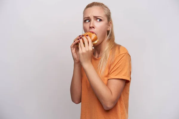 Portrait of confused young pretty blonde female with casual hairstyle looking thrillingly aside and rounding eyes while having lunch with fastfood, isolated over white background — Stock Photo, Image
