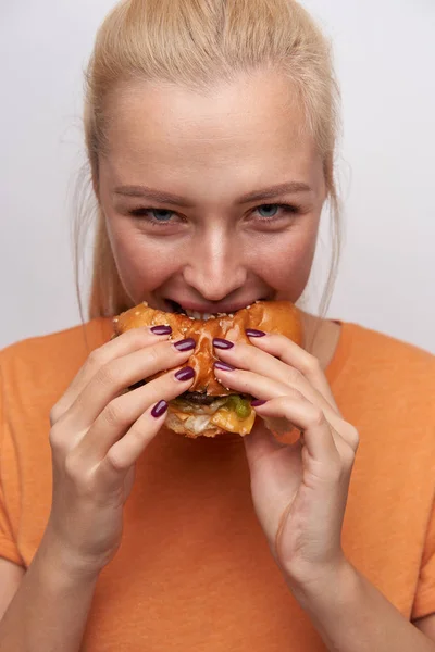 Vrolijk jong mooi blond vrouwtje met bordeaux manicure bijtend vraatzuchtig hamburger en vrolijk kijkend naar camera, gekleed in oranje t-shirt terwijl ze over witte achtergrond staat — Stockfoto