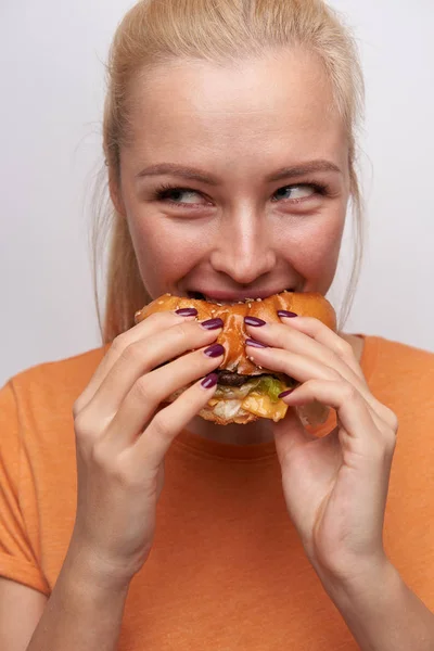 Close-up van verrukt jong vrij lang haar blonde vrouw met natuurlijke make-up genieten van haar hamburger tijdens het eten, vrolijk glimlachen en kijken opzij, geïsoleerd over witte achtergrond — Stockfoto