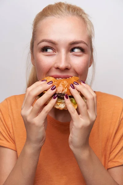 Positivo affamato giovane bella bionda signora con taglio di capelli casual guardando allegro a parte mentre mangia hamburger con grande piacere, in piedi su sfondo bianco in t-shirt arancione — Foto Stock