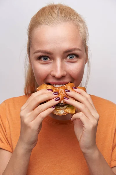 Porträt einer aufgeregten jungen blauäugigen hübschen blonden Frau, die eifrig ihren Burger isst und rundliche Augen hat, während sie freudig in die Kamera blickt und vor weißem Hintergrund in lässiger Kleidung posiert — Stockfoto