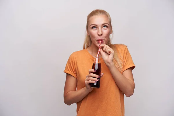 Una jovencita guapa rubia de ojos azules de largo pelo que bebe soda con paja y mira hacia arriba positivamente, lleva ropa casual mientras se apoya sobre fondo blanco. — Foto de Stock