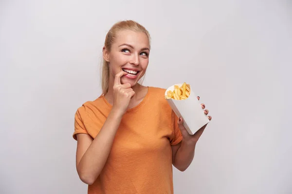 Alegre bonito de olhos azuis jovem loira fêmea segurando caixa de papel com batatas fritas e olhando para cima pensadamente com grande sorriso, contando calorias em sua mente, isolado sobre fundo branco — Fotografia de Stock