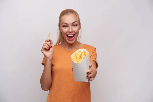 Studiofoto einer glücklichen attraktiven jungen blonden Frau, die sich über frische Pommes in der Hand freut und mit breitem Lächeln freudig in die Kamera blickt und vor weißem Hintergrund posiert — Stockfoto