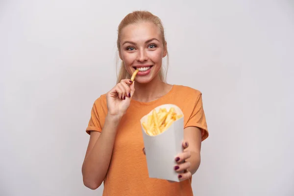 Joyful mooie jonge dame met lang blond haar kijken gelukkig op camera en ruiken breed tijdens het eten van frietjes, gekleed in casual kleding terwijl staan over witte achtergrond — Stockfoto