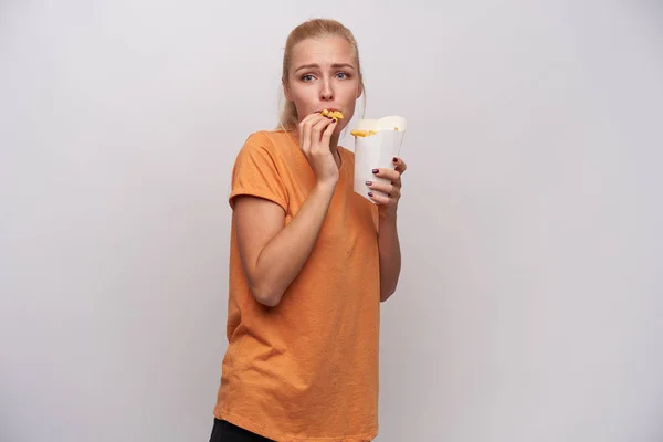 Emotionele jonge blauw-ogige blonde vrouw met paardenstaart kapsel kijken in de voorkant van haar en eten frites terwijl staan over witte achtergrond in oranje t-shirt — Stockfoto