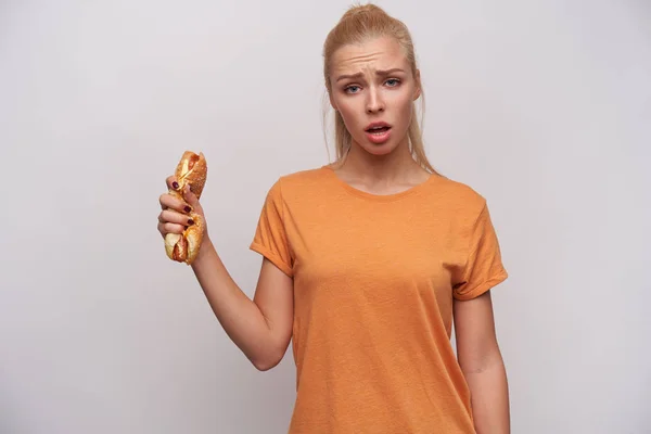 Ondeugende jonge langharige blonde vrouw met paardenstaart kapsel op zoek uitdagend naar camera en fronsende wenkbrauwen, het vasthouden van smashed hot dog in opgeheven hand terwijl staande over witte achtergrond — Stockfoto