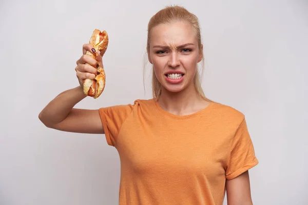 Indoor shot van boze jonge mooie blonde vrouw gekleed in casual kleding kijken gewelddadig op camera en verkreukelen hot dog in opgeheven hand, staande tegen een witte achtergrond — Stockfoto