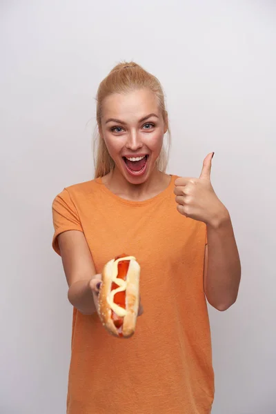 Studio photo of joyful young blue-eyed blonde lady with casual hairstyle showing hot dog in outstretched hand and raising thumb, looking happily at camera and smiling widely — Stock Photo, Image