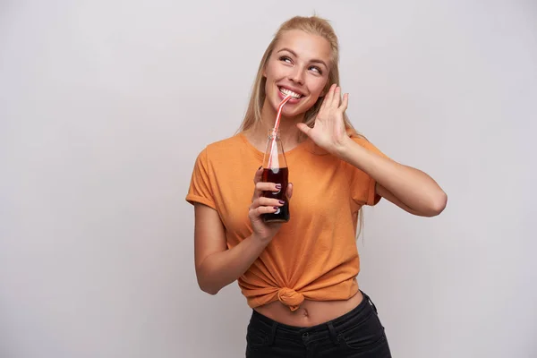 Foto interior de jovencita rubia rubia de pelo largo y alegre bebiendo soda y mirando alegremente a un lado con una sonrisa amplia, estando en buen estado de ánimo mientras se plantea sobre fondo blanco. — Foto de Stock