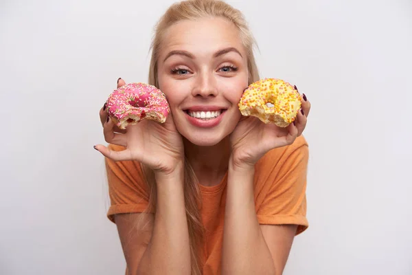 Cierre de la jovencita rubia guapa rubia que sonríe ampliamente en la cámara y que levanta sus amargas donaciones en la cara mientras se pone sobre fondo blanco con ropa casual. — Foto de Stock