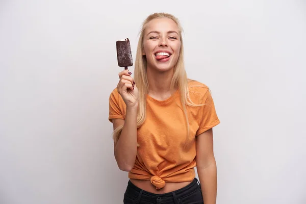 Estudio de tiro de una jovencita rubia alegre y atractiva con peinado casual sosteniendo crema de hielo en el palo y mostrando un lenguaje alegre en la cámara, de pie sobre fondo blanco. — Foto de Stock