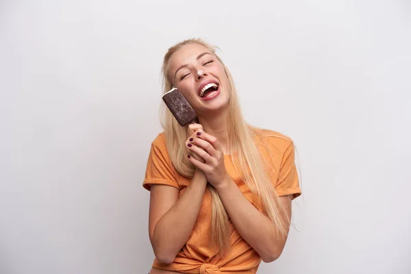 Studio foto van jong mooi uitziende langharige blonde dame in oranje t-shirt vrolijk lachend met gesloten ogen en knoeiend met ijs in haar handen, geïsoleerd over witte achtergrond — Stockfoto