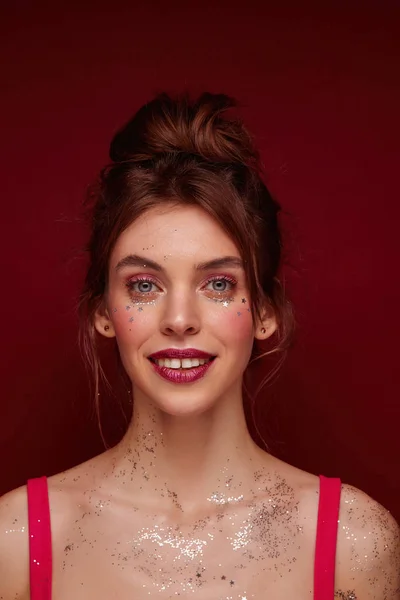 Portrait d'une belle jeune brune aux yeux bleus au maquillage festif et aux étoiles argentées sur le visage, debout sur un fond bourguignon, regardant positivement la caméra avec un sourire sincère Photos De Stock Libres De Droits