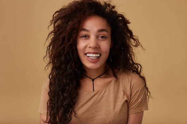 Portrait of happy young attractive brown haired curly woman with voluminous hairstyle showing her white perfect teeth while smiling gladly to camera, isolated over beige background