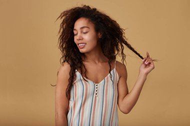 Hoş görünümlü, koyu tenli, esmer bir kadın. Gözleri kapalı gülümseyen, kıvırcık saçlarını kaldıran, bej arka planda izole bir kadın.