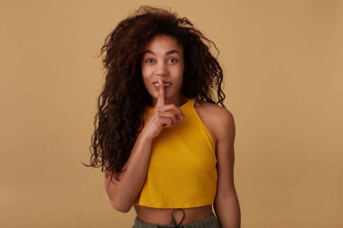 Pozitif, genç, kıvırcık saçlı, kıvırcık tenli, doğal makyajlı, işaret parmağını ağzına doğru kaldıran ve sırrını bej arka planda tutmak isteyen bir kadın.