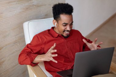 Siyah tenli, kısa saçlı, duygusal olarak elini kaldıran ve kaşlarını çatan, vido çağrısı yapan, evin içinde dizüstü bilgisayarla poz veren etkileyici genç adam.