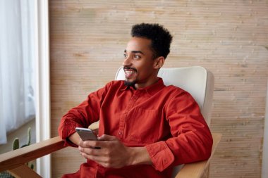 Genç, çekici, kısa saçlı, esmer, sakallı bir erkeğin portresi. Koyu tenli, elinde akıllı bir telefon, neşeli bir şekilde pencereden dışarı bakıyor. Evin iç kısımlarına bakıyor.