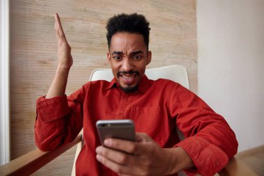 Meşgul, kahverengi gözlü, kısa saçlı, koyu tenli, cep telefonundan kafasını karıştıran ve ev içi koltukta otururken elini kaldıran adam.