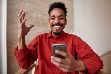 Yakışıklı, kısa saçlı, koyu tenli, gözlerini kapalı tutan, mutlu bir şekilde gülen ve elini kaldırmış, bej rengi iç kısımda izole bir şekilde cep telefonu tutan genç bir adam.