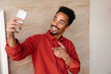 Genç, çekici, kısa saçlı, kıvırcık saçlı, esmer, esmer erkek. Elini kaldırarak selfie çekiyor ve kameraya bakarak bej rengi iç kısımda poz veriyor.