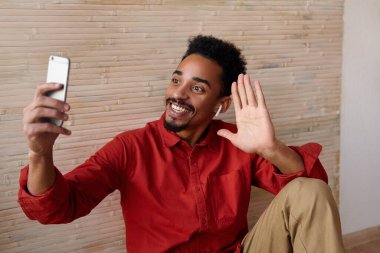 Kapalı alanda kısa saçlı, koyu tenli, el kaldıran genç bir erkeğin fotoğrafı. Merhaba jesti ve neşeli bir şekilde gülümsüyor.